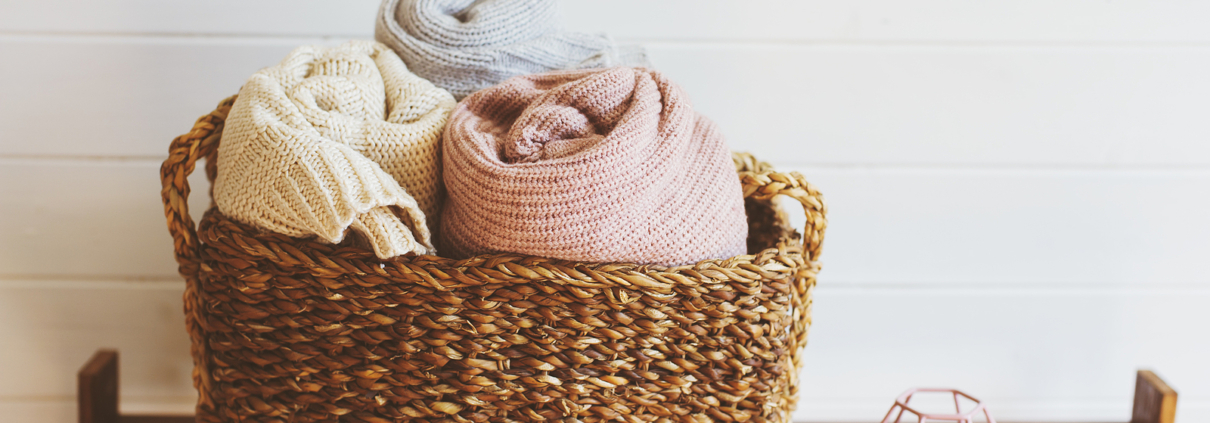 Image of an organized towel basket. In a brown wicker basket are three towels that are rolled. 1 light yellow, 1 baby blue, and one baby pink. The basket is on top of a wooden shelving unit. Only the top shelf is in frame. Beside the basket is a small tea candle in a metal geometric holder. The wall behind the basket is white.