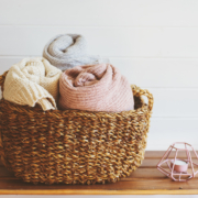 Image of an organized towel basket. In a brown wicker basket are three towels that are rolled. 1 light yellow, 1 baby blue, and one baby pink. The basket is on top of a wooden shelving unit. Only the top shelf is in frame. Beside the basket is a small tea candle in a metal geometric holder. The wall behind the basket is white.